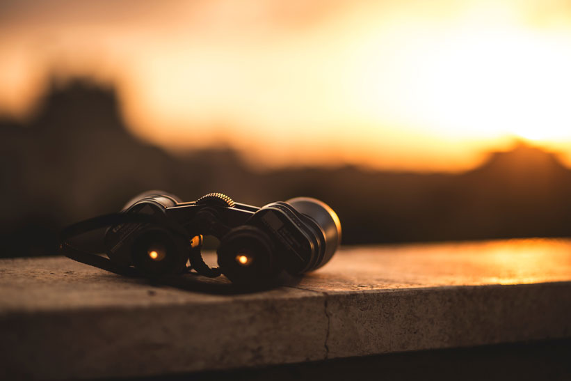 A photo of a pair of black binoculars sitting on a concrete ledge with a blurry sunset in the background