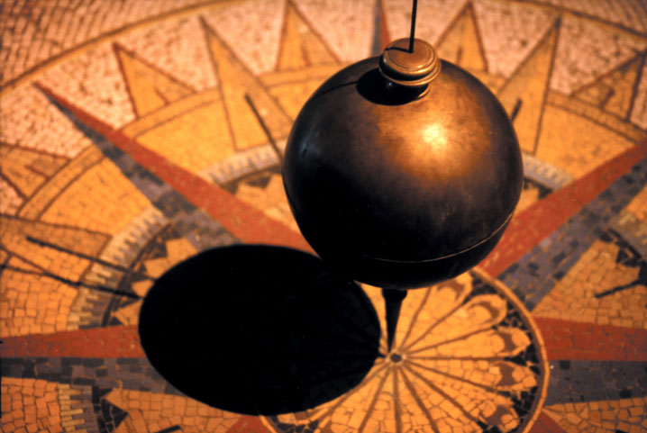 A photo of a brass pendulum suspended over a mosaic floor in the pattern of a sunburst, with a dark shadow cast by the pendulum