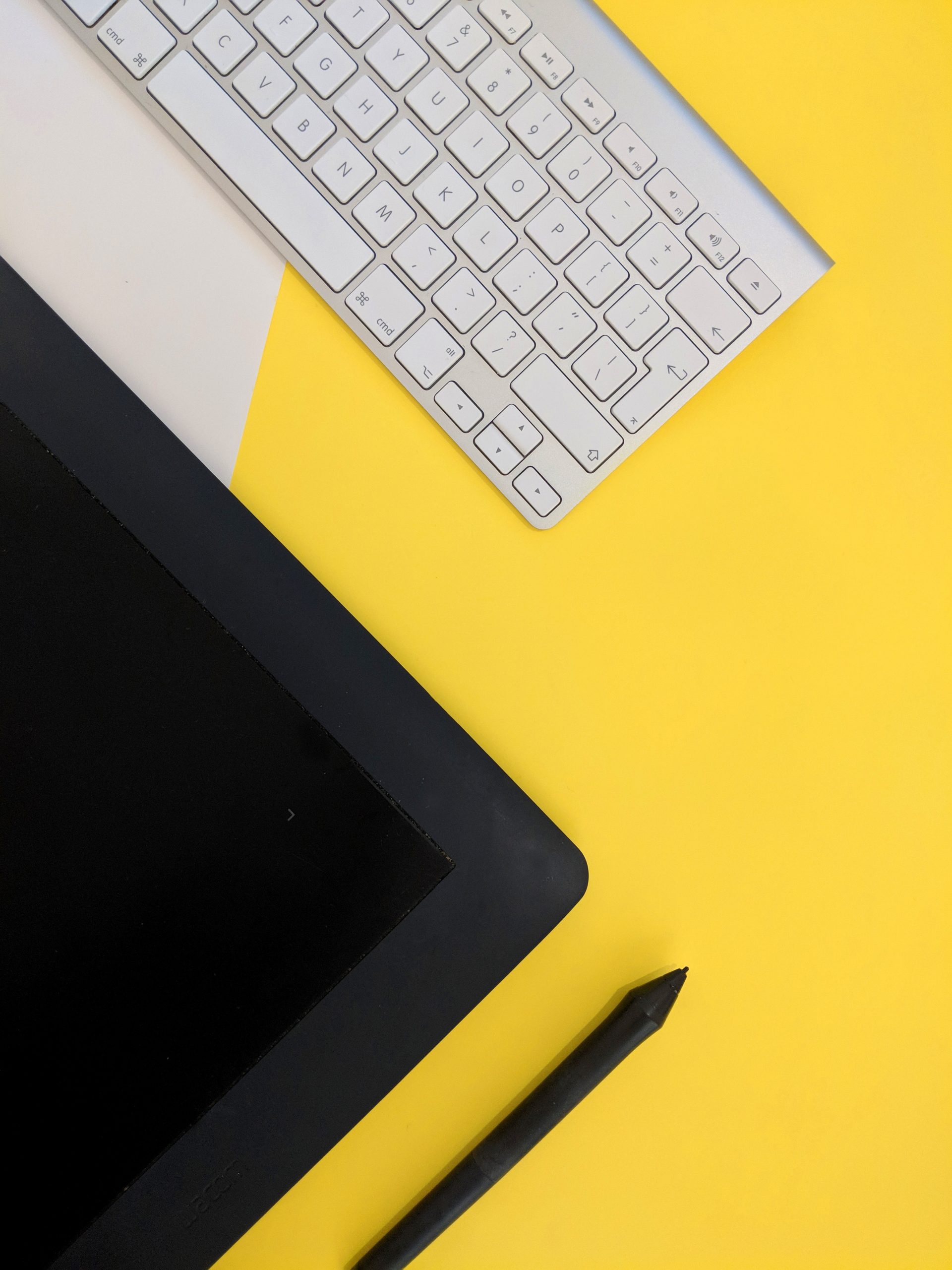 A photo of a silver Mac keyboard and a black tablet and stylus against a bright yellow and white background