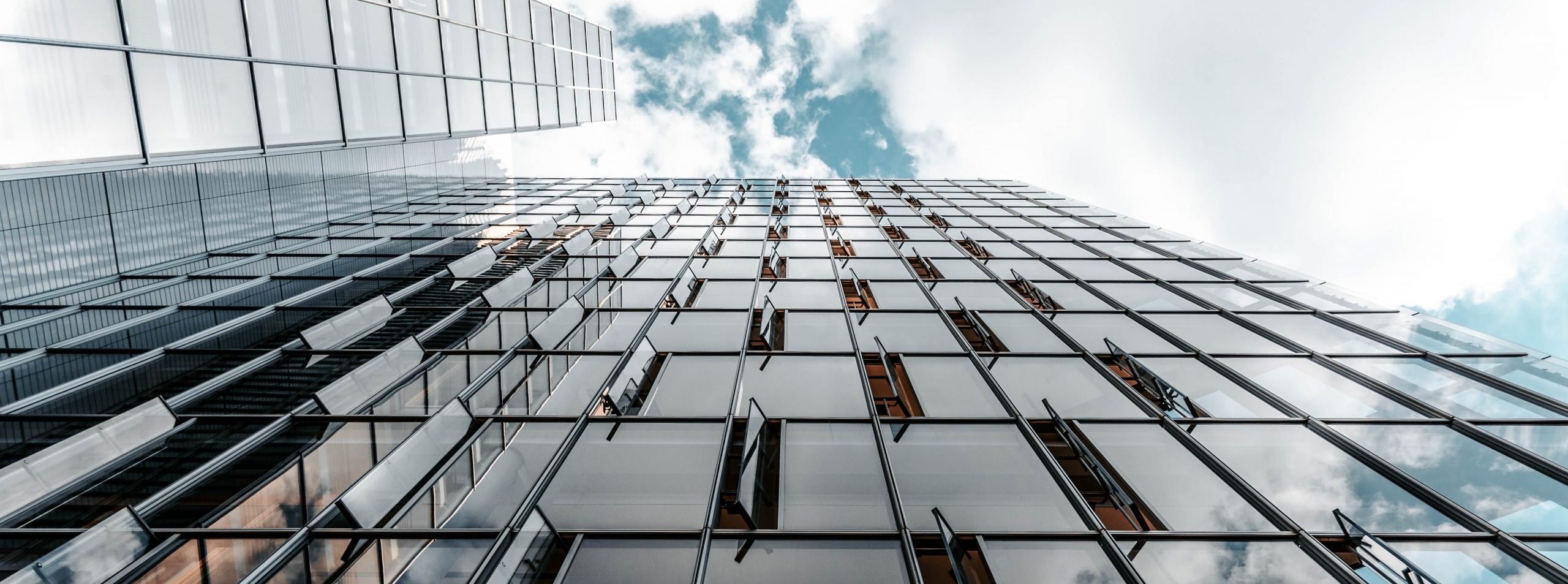 A low-angle photo of a glass building with some open windows against a cloudy blue sky