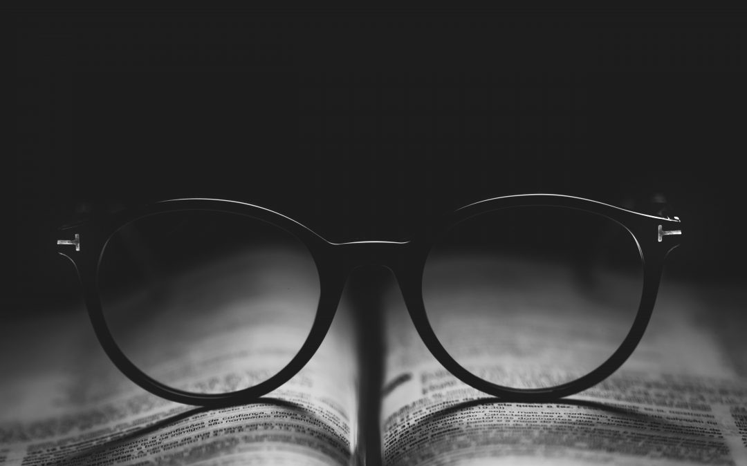 A black and white photo that depicts a pair of glasses sitting on the open pages of a book.